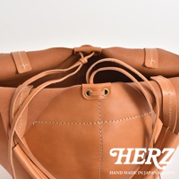 職人手作皮革球形肩掛托特包，呈現自然棕褐色，採用柔軟皮革材質，以白色縫線勾勒出包身線條，並以皮革繫帶束口，整體散發簡約質感。