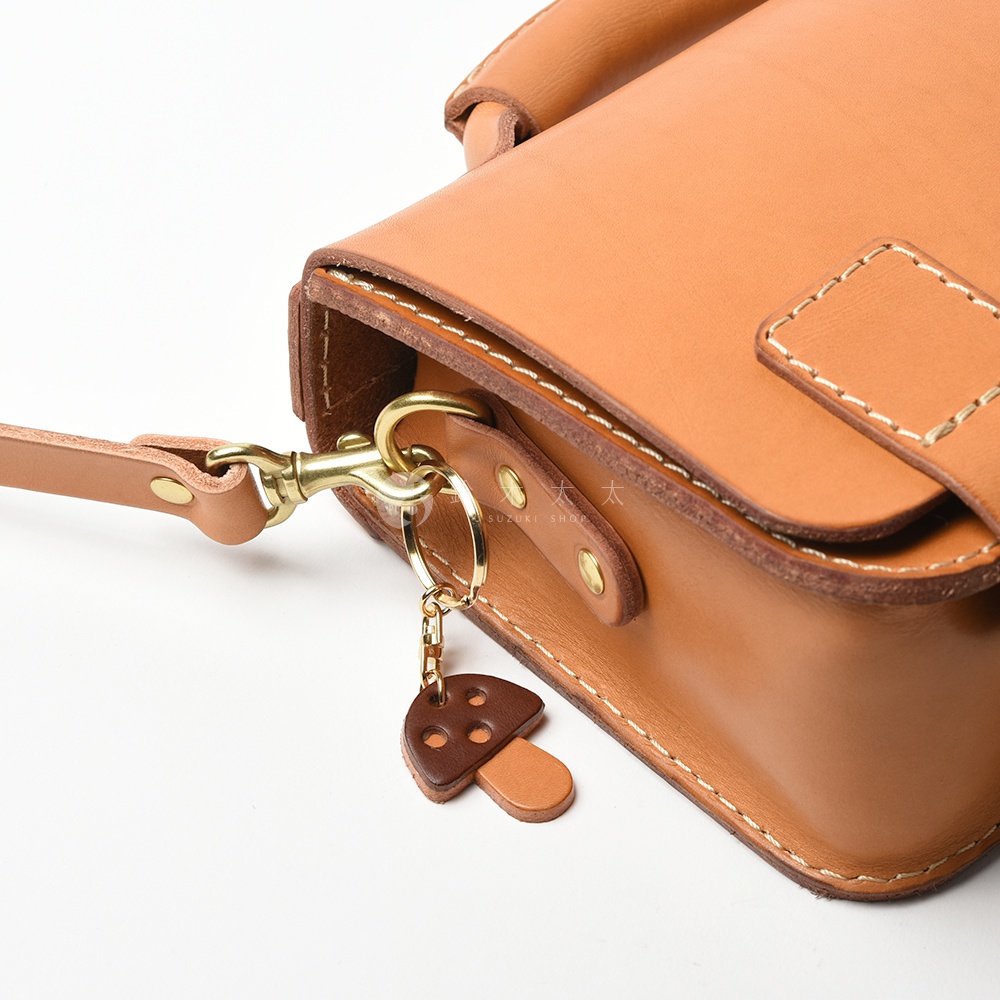 職人手作皮革香菇造型鑰匙圈，採用淺棕色皮革製作，香菇傘部為深棕色，傘下有四個小圓孔裝飾，搭配金色金屬掛環與鉤扣。