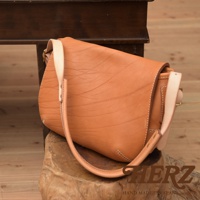 原色皮革製作的郵差包，帶有自然的皺褶紋理，搭配淺色皮革的背帶，包身為圓弧造型，以白色縫線點綴，呈現簡約復古風格。