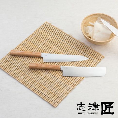 兩把Yuri / Santoku萬用刀，刀刃為銀色不鏽鋼，刀柄為淺木色，放置於竹製墊上，旁邊有一個裝有切片食材的木碗。