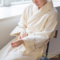 一位女性穿著米白色今治純棉浴袍，繫著腰帶，坐在椅子上，浴袍領口和袖口有細緻的滾邊裝飾，材質看起來柔軟舒適。