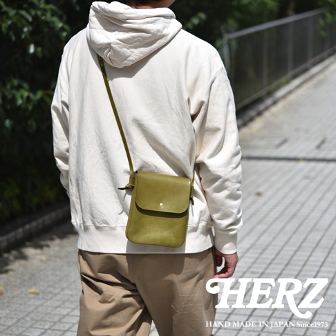 一位背著日本HERZ手作橄欖綠色皮革隨行袋的男子，他身穿白色連帽上衣與卡其色長褲，斜背的皮革包款線條簡約，正下方標示著HERZ品牌字樣與創業年份。