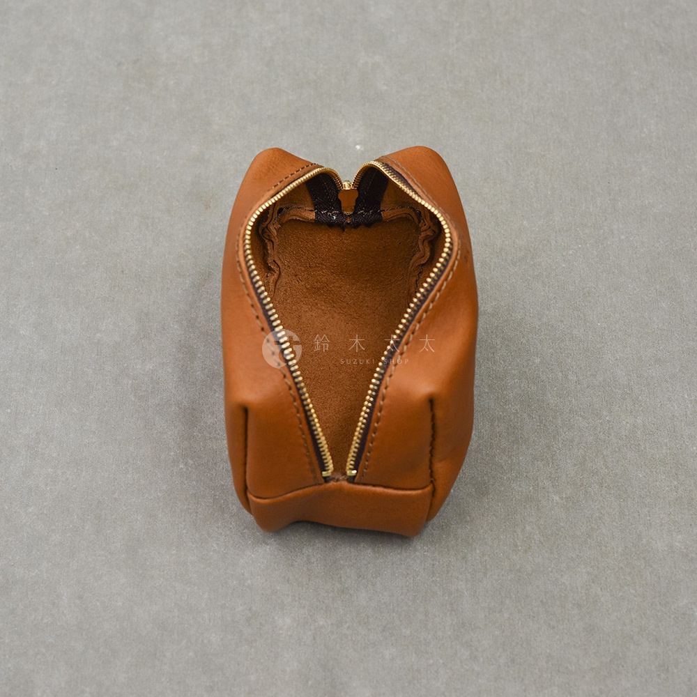 這是一個頂視角拍攝的日本HERZ職人手作皮革拉鍊收納包，整體呈現溫潤的焦糖棕色。皮革表面具備自然紋理與質感，包身採用直立式設計，中間由金屬拉鍊開合，內部呈現原始的麂皮絨面質感，整體簡約且帶有精緻的手工縫線細節，背景為中性灰色調。