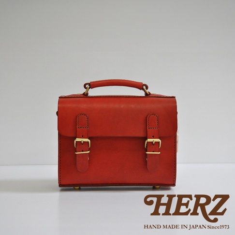 紅色皮革手提包，帶有兩個金屬扣環的翻蓋設計，頂部有提把，整體呈現簡約風格。