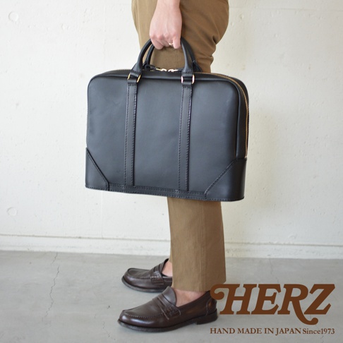 黑色皮革公事包，配有金色拉鍊和把手。包包設計簡潔，有明顯的縫線細節，風格經典。