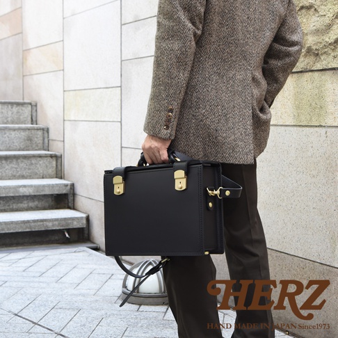 黑色皮革製作的職人手作兩用機師箱，箱體方正，配有金色鎖扣及提把，可肩背或手提。
