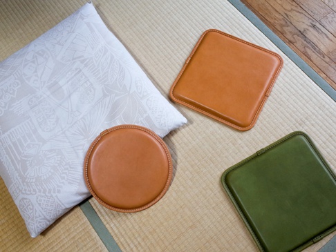 職人手作皮革圓形坐墊，呈現溫潤的棕褐色，皮革表面光滑，車縫線精緻。旁邊有一個同色系的方形皮革坐墊，以及一個綠色的方形皮革坐墊，均放置在榻榻米墊上。