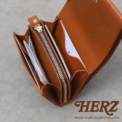 這是一款日本職人手工製作的HERZ品牌Organ系列皮革短夾，呈現焦糖棕色澤，質感紮實細膩。圖片採高角度俯視，展示皮夾展開後的內部構造，包含中間的拉鍊式零錢袋、兩側摺邊的卡片收納隔層，以及高品質的金屬拉鍊細節，右下角印有HERZ品牌標誌與手工製造字樣。