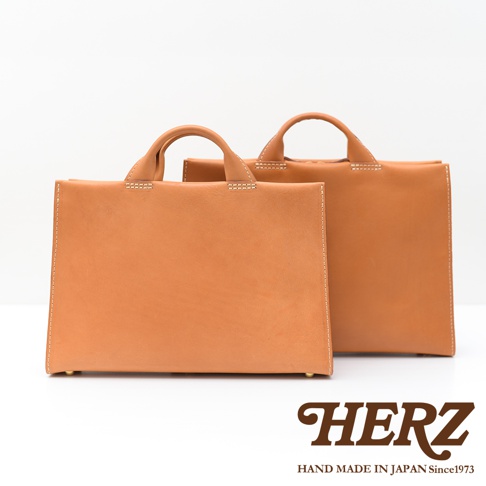 一前一後擺放著兩個日本製手工皮革包，前方的包包較大，呈現簡約的方形設計，揹帶為手提式；後方的包包較小，同樣為方形，揹帶也是手提式。兩款包包的皮革顏色均為淺棕色，表面有清晰的縫線細節，底部有金屬腳釘。圖片下方有HERZ的品牌標誌和「HAND MADE IN JAPAN Since1973」的文字。