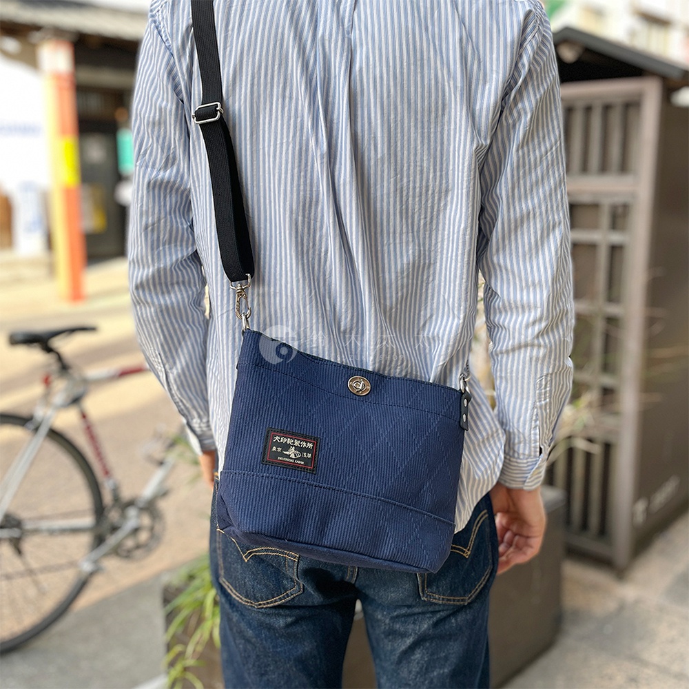 深藍色刺子織迷你側背包，表面有菱格紋縫線，正面有一個帶有日文標誌的黑色皮牌，配有可調節的黑色肩帶。