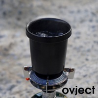 黑色琺瑯馬克杯，杯內有正在沸騰的水，置於戶外露營爐具上，背景為模糊的地面。