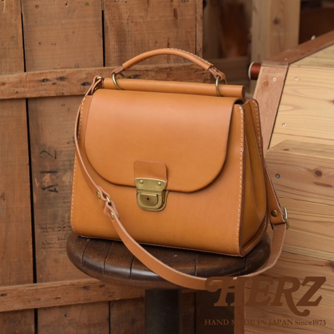 日本HERZ職人手作駝色皮質兩用包，配備金屬鎖扣與可調式背帶，置於木製背景上。