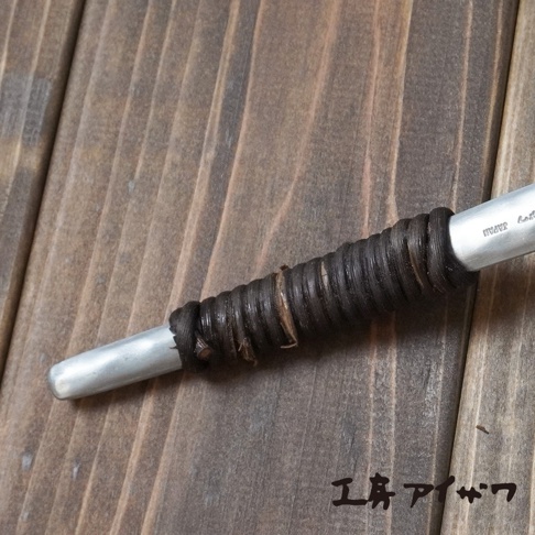 銀色鋁製湯勺，勺柄部分纏繞著深棕色繩狀材質，繩狀材質有明顯的紋路和部分乾枯的植物纖維，勺柄末端刻有「JAPAN」字樣。