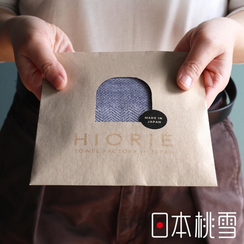 雙手捧著一包裝著今治蓬鬆人字紋紗布方巾的牛皮紙袋，上面印有HIORIE TO WEL FACTORY in JAPAN字樣，並有MADE IN JAPAN的圓形標籤。