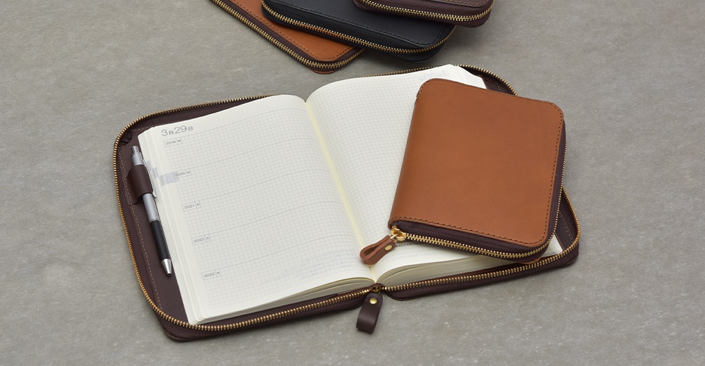 棕色皮革記事本收納包打開，露出內部網格狀的空白內頁，一支筆夾在左頁。