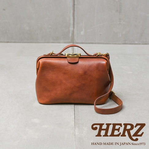 日本HERZ職人手作皮革Organ鎖孔式大開口肩背包，展現自然紋理的棕色皮革，搭配復古鎖孔設計，背帶自然垂落，背景為灰色水泥牆。