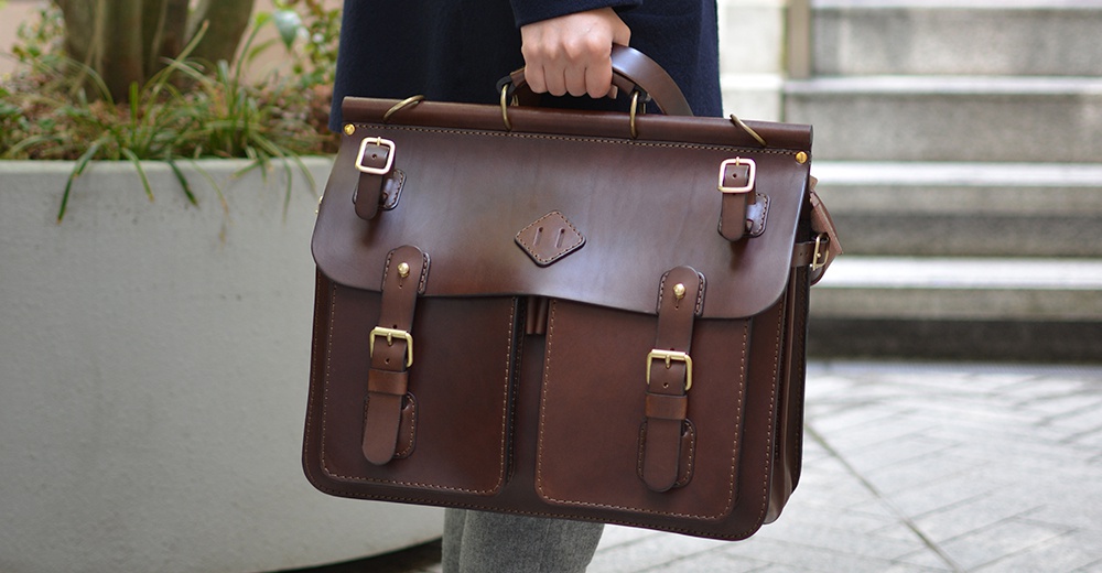 職人手作皮革旅行包 A-102，深棕色，採用光滑皮革製作，配有黃銅色金屬扣環和提把，包身有兩個前袋，設計簡約實用。