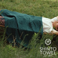 一塊深綠色的2.5層紗材質隨身暖毯，蓋在躺在戶外的人身上，毯子呈現自然的皺褶感，質感柔軟。