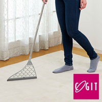 一隻手握著銀色金屬掃把，掃把頭為灰色，扇形設計，帶有圓形镂空圖案，底部有灰色膠條。掃帚置於淺米色地毯上，一位穿著深藍色牛仔褲和灰色條紋襪子的人站在右側。