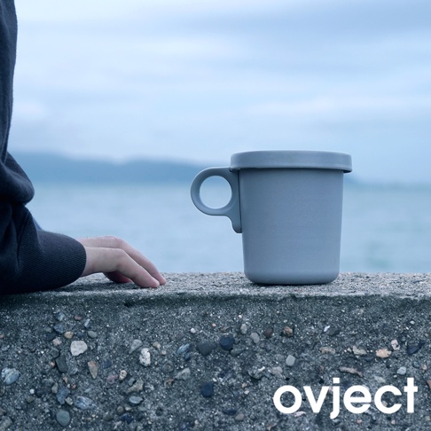 灰色琺瑯馬克杯，配有圓形把手和密封杯蓋，置於戶外石牆上，背景為海景與天空。