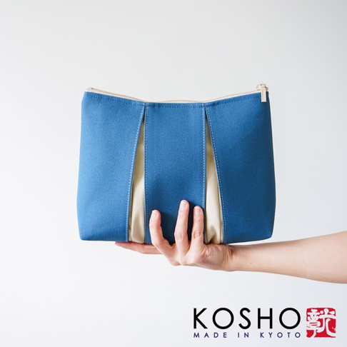 一隻手托著京都KOSHO品牌的ougi帆布化妝收納包，該收納包為藍色與米色拼接設計，外觀呈現簡約的立體折疊造型，右下角印有KOSHO品牌名稱及紅色日文印章標誌。