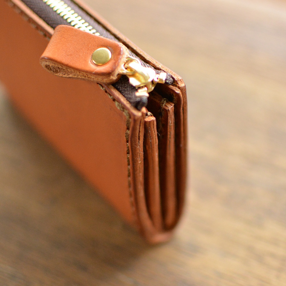 焦糖色皮革短夾，特寫拉鍊細節，帶有金色金屬拉頭與按扣，皮革質感紋理清晰，置於木紋背景上。
