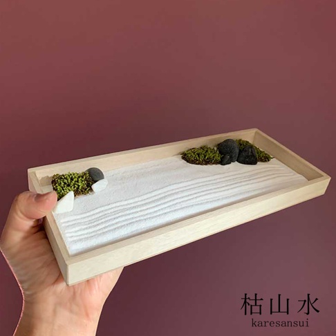 淺色木製托盤內，白砂被耙出層層漣漪，點綴著幾叢綠色苔蘚、灰黑色的圓石與白色小石塊，呈現日式枯山水庭園景觀。