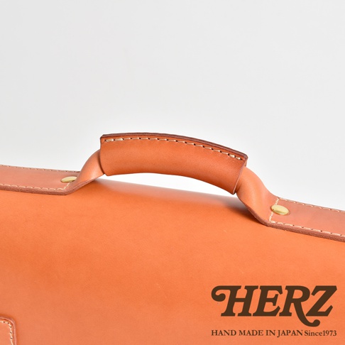 職人手作皮革經典三扣兩用商務公事包，特寫光滑的淺棕色皮革手提把，帶有精緻縫線與鉚釘固定，包身為溫潤的橘棕色調。