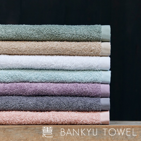 疊放著八條長纖維浴巾，顏色由上至下分別為：薄荷綠、米褐、白色、淺藍綠、薰衣草紫、灰藍、淺粉紅，以及最底部的淡粉色。浴巾材質看起來柔軟蓬鬆，邊緣有精緻的織邊。