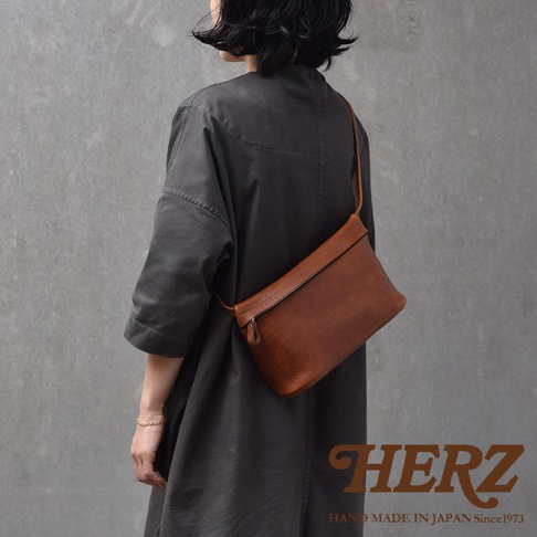 一名女性身穿深灰色長袍，側身展示一個棕色皮革斜背包，包包橫跨在胸前。