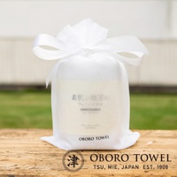 白色束口袋裝著一條印有日文「素肌、微笑」與英文「bare skin smiles FACE TOWEL」的毛巾，束口袋頂部綁著白色緞帶，放置於木質桌面，背景為模糊的綠色草地與建築。