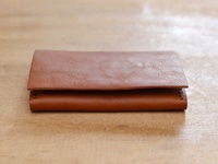 一個棕褐色的薄型皮革名片夾，表面有自然紋理，採用手工縫線工藝，置於木紋背景上。