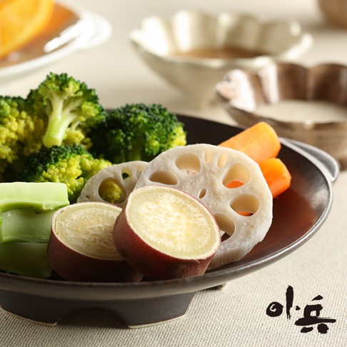 深棕色美濃燒蒸盤盛著蒸熟的蔬菜，包含綠色花椰菜、切片蓮藕、紅皮黃肉的地瓜和胡蘿蔔塊。