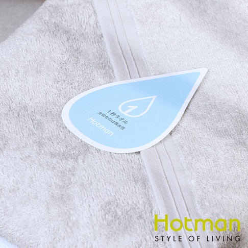 一條淺灰色毛巾，表面柔軟細緻，靠近中間處有一張藍色水滴形狀的標籤，上面印有白色數字「1」和日文說明，下方有「Hotman」品牌字樣。