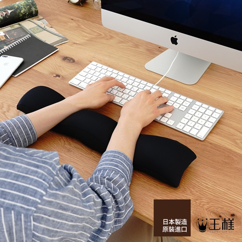 黑色鍵盤減壓護腕枕，柔軟材質，符合人體工學設計，支撐使用者手腕，搭配白色蘋果鍵盤與電腦螢幕，擺放在木質桌面。