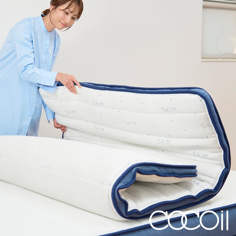 一位女士正在展示一張可折疊的雙層獨立筒床墊。床墊表面為白色，印有藍色「SKIN」字樣和十字圖案，邊緣為藍色滾邊。