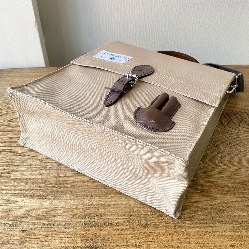 犬印純綿帆布三用直式側背包，淺卡其色，具有兩個皮質束帶扣環，一個用於固定翻蓋，另一個為裝飾性設計，帆布材質，簡約實用。