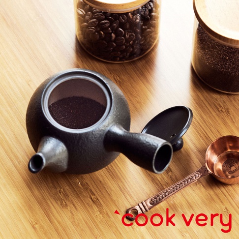 黑色常滑燒咖啡急須茶壺，壺身呈圓潤的球狀，帶有短嘴和提把，壺內裝有咖啡粉。旁為銅製咖啡量匙及玻璃罐裝咖啡豆。