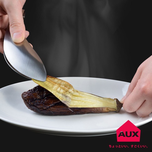 銀色金屬材質的調理夾，夾著一塊正在被剝皮的茄子，茄子呈現深棕色與淡黃色，放在白色盤子上。