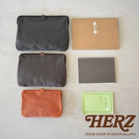 職人手作皮革Organ手拿包，展示黑色、咖啡色與橘色三款。包款為簡約設計，帶有金屬扣環，材質為皮革。一旁配有牛皮紙信封、灰色筆記本及綠色筆記本。