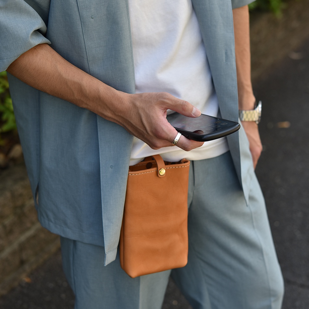 棕褐色皮革製作的長方形手機包，附有黃銅色按扣和提把，懸掛於穿著藍灰色套裝的人腰間。