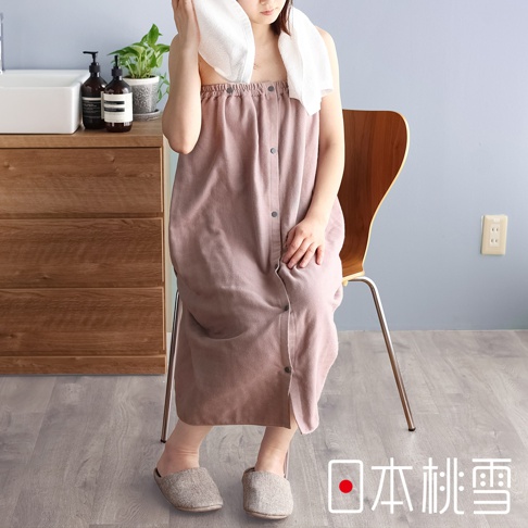 一位女性穿著一件淡粉色的棉紗混紡平口鬆緊浴裙，浴裙有按扣設計，她坐在木質椅子上，頭上搭著白色毛巾。