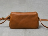 職人手作的棕色皮革輕巧拉鍊單肩包，設有金色拉鍊和可調節的皮革肩帶，包身呈現方形的長條狀。