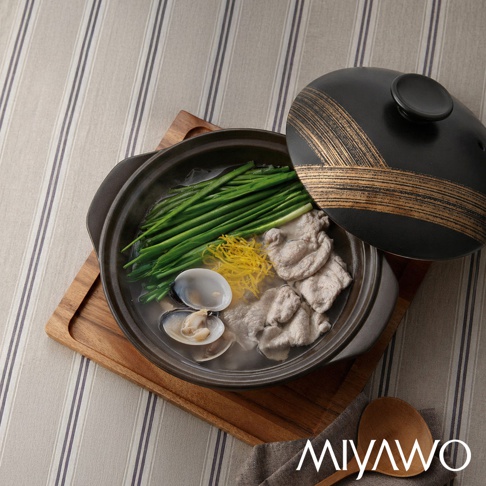 一個黑色宮尾MIYAWO陶瓷湯鍋，半開的鍋蓋帶有金色筆觸紋理，鍋內盛裝著新鮮的蛤蜊、翠綠韭菜、薄切肉片與黃色蛋絲，整鍋置於木質托盤上，背景為簡約的灰色條紋桌布。