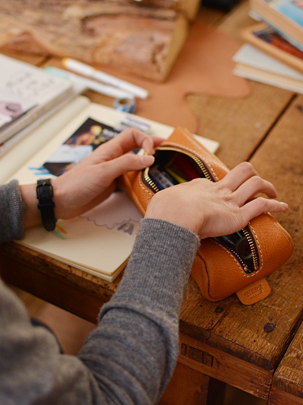 職人手作皮革圓角收納筆袋，棕色皮革，拉鍊開合，內有彩色筆，搭配灰色毛衣，置於木桌上，旁有筆記本及書籍。