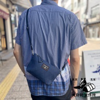 藍色刺子織隨身斜背包，方形格紋縫線，配有黑色斜揹帶和金屬扣環，包上貼有紅色與白色標籤，由一位身穿藍白格紋襯衫的男士斜揹於身。