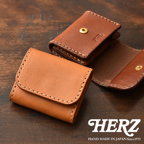 兩款日本HERZ職人手作皮革零錢包置於木質桌面上，前方為淺棕色皮革零錢包，後方為深棕色皮革零錢包，其設計簡約並以耐用的粗線車縫，上方壓印有HERZ品牌字樣，展現細膩的手工質感。