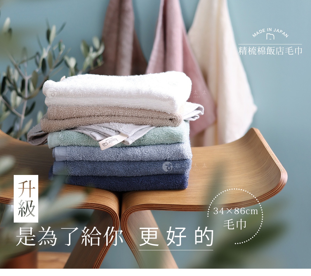 圖片文字：日本製精梳棉飯店毛巾，尺寸為三十四乘以八十六公分。畫面中一疊多種顏色的毛巾堆疊在木製弧形椅上，後方懸掛著幾條同系列毛巾，整體呈現溫馨舒適的質感。升級是為了給你更好的，強調該產品的高品質與舒適體驗。