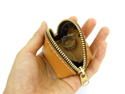 手持的職人手作皮革貝殼造型零錢包，顏色為自然焦糖色，材質為真皮。拉鍊為金色，內部可見兩個戒指。