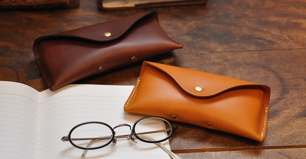 兩款職人手作皮革眼鏡收納盒，分別為深棕色與淺棕色，皆以精緻車縫工藝製作，搭配金屬按扣，置於木質桌面與筆記本旁。